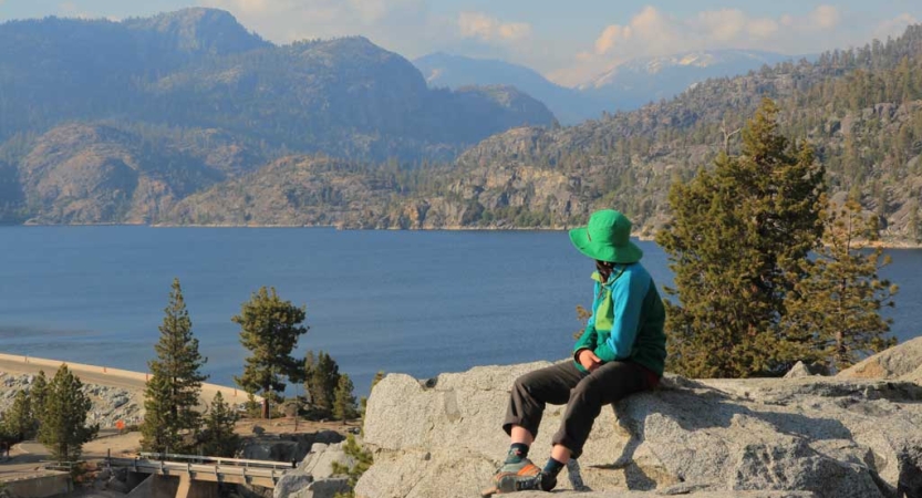 A person sits on a rock, looking out over a lake that is framed by mountains. 